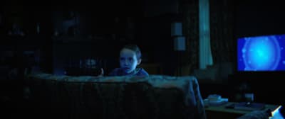 En liten flicka tittar upp bakom en soffa i ett mörkt rum med skenet från en tv-ruta i bakgrunden.