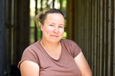 En kvinna med ljust hår och iklädd brun t-skjorta ser in i kameran och ler. I bakgrunden en träpanel.