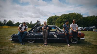 Matha lutar sig mot en svart, amerikansk bil och röker, bredvid henne står och sitter tre män. Bilen är parkerad på en äng, i bakgrunden syns träd och en blå himmel.