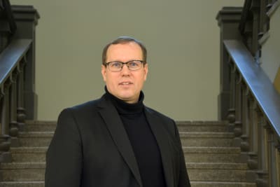 Vit man i 40-årsåldern med glasögon, svart polo och svart kavaj, står i en trappa och tittar rakt in i kameran.