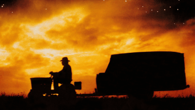 Oranssina hehkuvaa taivasta vasten näkyy mies, joka istuu ajettavan ruohonleikkurin päällä ja vetää perässään kuormaa, kuva elokuvasta The Straight Story.