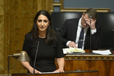 Nasima Razmyar (SDP) talar i riksdagens plenum. Bakom henne skymtar statsminister Petteri Orpo (Saml) i regeringsbåset. Han håller sig för pannan.