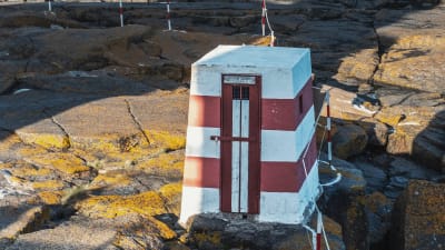 Litet, rödvitrandigt, fyrkantigt hus som fungerat som utedass på klippa.
