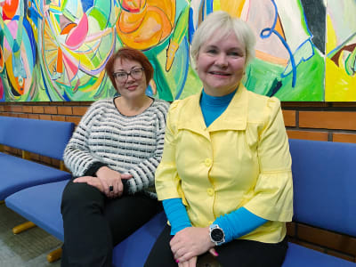 Olga Biriukova och Maryna Vedmid sitter bredvid varandra framför mural på Helsingfors arbis.