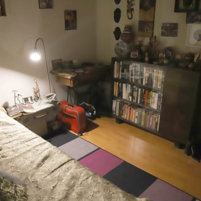 Ett ganska mörkt gästrum med säng, bokhylla, golvlampa som lyser och en kappsäck.