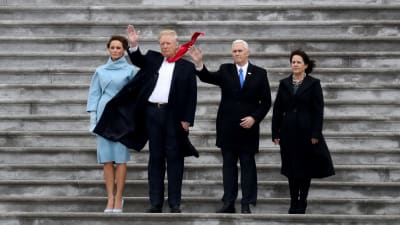 Donald trump, Melania Trum, Mike Pence med fru står i blåsten och hälsar på folkmassan. Trumps röda slips fladdrar i vinden.