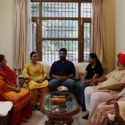 Intialainen kuusihenkinen perhe istuu sohvilla. Kuvassa on kaksi miestä ja neljä naista. Vanhemmalla miehellä on päässään punainen turbaani.