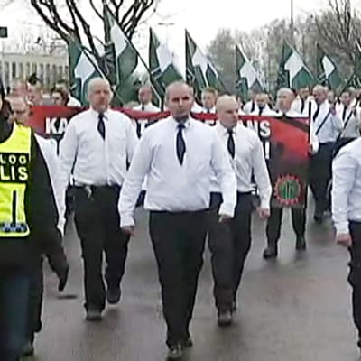 Valkopaitaisten ja mustakravattisten miesten rivistö kantaa vihreä-valkoisia lippuja. Edessä kulkee poliisi. 