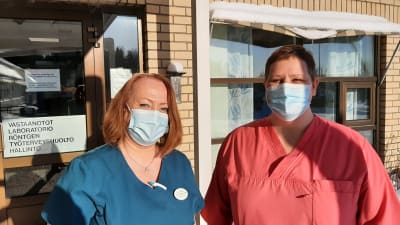 Två hälsovårdare med munskydd vid ingången till en hälsocentral, Carola Silander-Lehtinen i blå sköterskedräkt och Camilla Rautanen i röd sköterskedräkt.