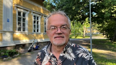Man med glasögon framför trähus. 