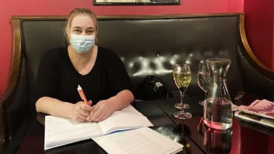 En kvinna sitter vid ett bord, och arbetar med nåt slags manuskript. Hon har på sig munskydd. På bordet står vinglas.