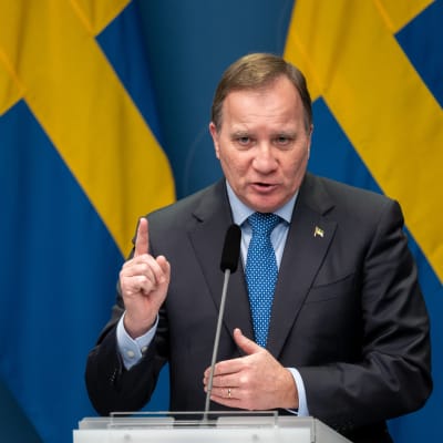 Stefan Löfven på pressträff framför svenska flaggor, med pekfingret höjt i förmaning.