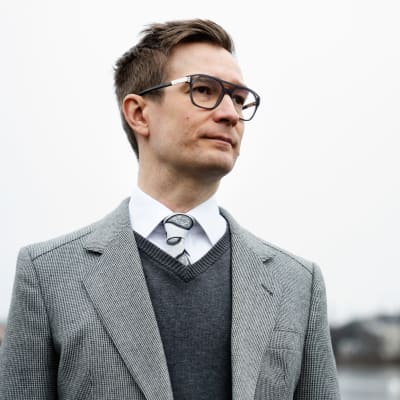 Silmälasipäinen mies katsoo kaukaisuuteen ja hänen takanaan on vettä, rantaa ja taivasta.