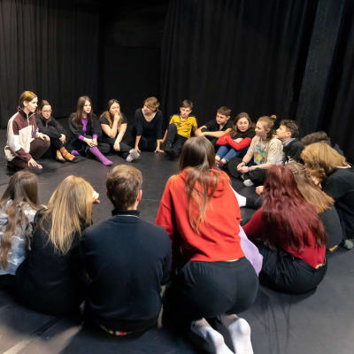 Ukrainalainen lasten- ja nuortenteatteriryhmä istuu ringissä Huoneteatterin lavalla ja leikkii teatterileikkiä. Ryhmässä on parikymmentä jäsentä.