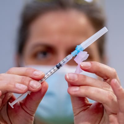 En kvinna med munskydd håller en spruta med vaccin framför sitt ansikte.