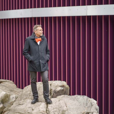 Tapio Vanajas seisoo kalliolla, taustana tummanpunainen ritiläseinä.