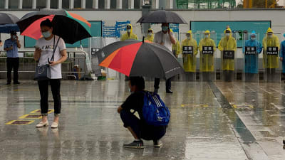 I förgrunden några personer med paraply, och i bakgrunden en rad personer i regnrockar och utrustade med sköldar med texten "polis".
