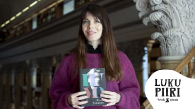 Kirjailija Riikka Pulkkinen Kansalliskirjaston antiikin pylvään vieressä Lumo-kirja kädessään.