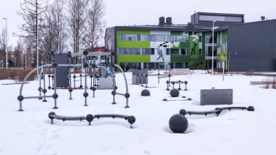 Olika ställningar och gymredskap i snön utanför en grön byggnad.