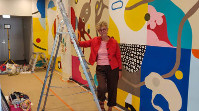 En kvinna i rött lutar sig mot en metallstege. Bakom henne syns en färggrann abstrakt väggmålning.
