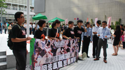 En rad demonstranter står bakom en banderoll på ett torg i Hongkong