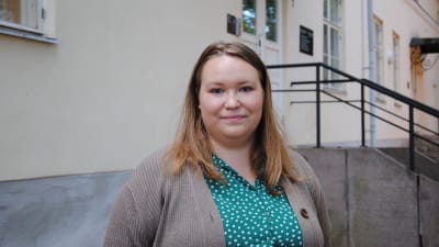 Kvinna med blont hår, och gråbrun stickad tröja står utanför byggnad.