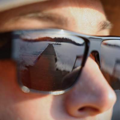 En man i solglasögo, som reflekterar en bild av en sjö och en brygga.