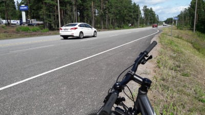 Bilar kör på en riksväg. I förgrunden syns ett cykelstyre.