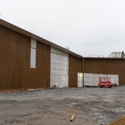 En byggarbetsplats fotad utifrån. Arbetsmaskiner står parkerade utanför huset och plast täcker områdena i fasaden där det ska finnas fönster.