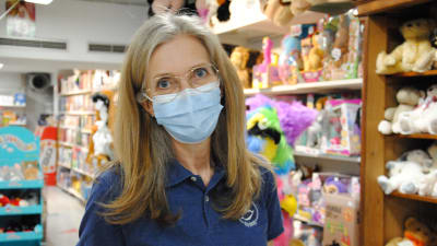 En kvinna med ljust hår, tunna glasögon och ansiktsmask står inne i en leksaksaffär.