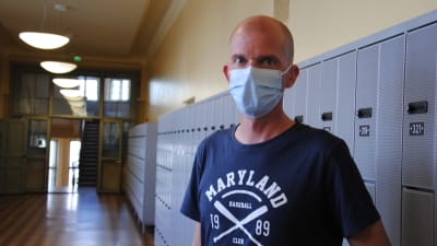 Skallig man med mörkblå t-skjorta med tryck och munskydd står framför skåp i en skolhall.