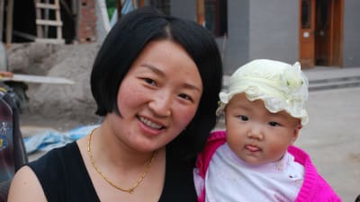 En kinesisk mamma och hennes dotter. Mamman har page, svart topp och ett guldsmycke. Flickan är ännu ett spädbarn och klädd i en ljusgul hatt. Hon sträcker ut tungan.