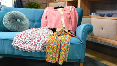 En blommig kjol, en rosa jacka och en blommig klänning som är sydda av återvunna tyger. Plaggen ligger framlagda på en blå sammetssoffa.