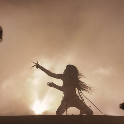 En kvinna med långt, mörkt hår framför en sång på en scen. Hon är fotograferad i motljus.