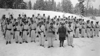 Lumipukuun pukeutuneita sotilaita rivissä aseet olalla talvella