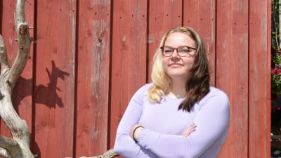 Ung kvinna i lilafärgad tröja poserar framför ett rött trähus en varm sommardag.