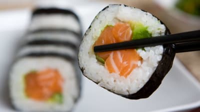 En bit rullad maki-sushi mellan två matpinnar med flera sushibitar i bakgrunden.