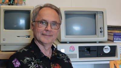 Porträtt på it-företagaren Petteri Järvinen sittande i blommig skjorta framför två datorer från 1980-talet, en IBM PC/XT och en Apple Macintosh Plus. I bakgrunden även exemplar av Järvinens klassiska bok PC-käyttäjän käsikirja (PC-användarens handbok).