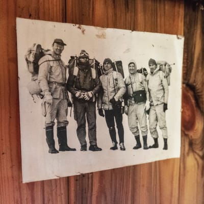 Svartvitt fotografi på trävägg. Bilden föreställer fem män med ryggsäckar och varma kläder på väg på expedition på Grönland.