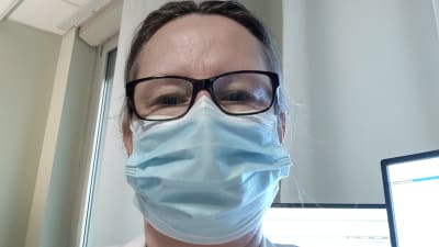 En kvinna med glasögon, läkarrock och munskydd tar en selfie.