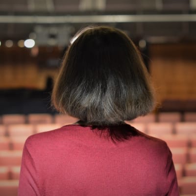 En kvinna i brun page och röd tröja ser ut över en tom teatersalong.