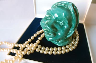 Ett grön skulptur föreställande ett kvinnohuvud med ett pärlhalsband i ett smyckeskrin. 