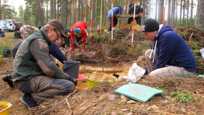 Flera personer sitter på huk vid ett arkeologiskt utgrävningsområde.