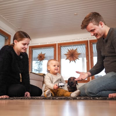 Vanhemmat, vauva ja mäyräkoira istuvat olohuoneen lattialla leikkimässä.