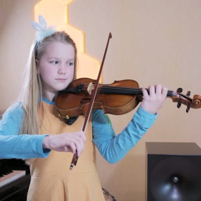 Keltaiseen mekkoon ja turkoosiin paitaan pukeutunut tyttö soittaa viulua. Taustalla näkyvät pianon koskettimet.