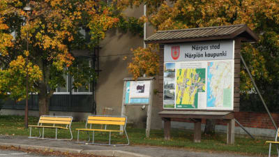 En infoskylt med kartor över Närpes. Till vänster om skylten två gula parkbänkar.