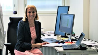 En kvinna iklädd klänning och kavaj, sitter vid ett skrivbord med många dataskärmar. Hon är en forskare. 