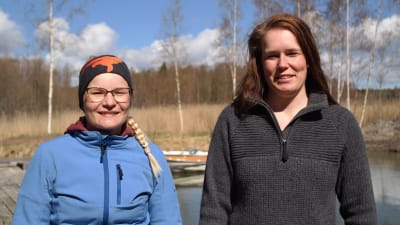 Sandra Bergqvist och Linda Sandström står på en strand med vatten, björkar och vass i bakgrunden
