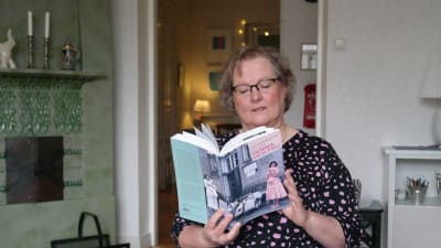 Lena Marander-Eklund sitter framför en kakelugn och läser ur boken Hemma och fru