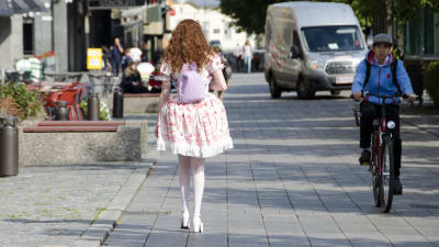 En kvinna klädd i så kallade Lolitakläder - en fluffig vit och rosa klänning med spetsar på. Hon har vita högklackade skor, blommor i håret och en lila ryggsäck med kaninöron på. Vi ser henne bakifrån när hon går på en trottoar i centrum av Vasa. 